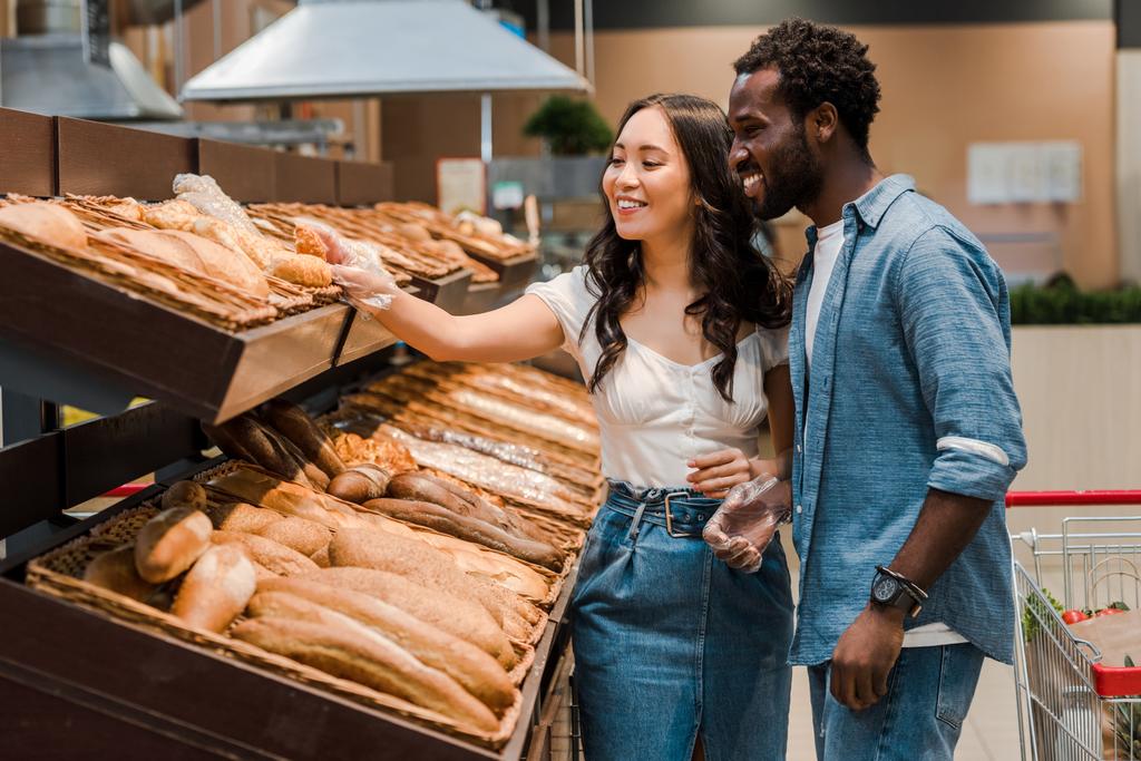 boldog ázsiai nő megható kenyér állva közel afro-amerikai ember a szupermarketben  - Fotó, kép