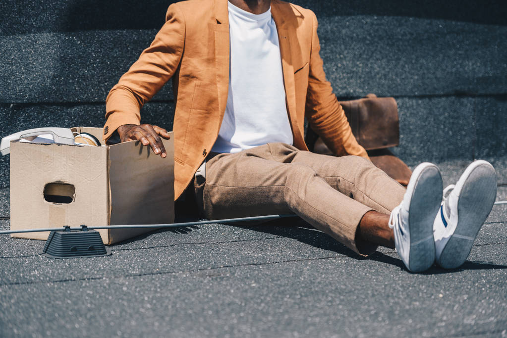 vista recortada de hombre de negocios afroamericano despedido sentado en la azotea cerca de caja de cartón
 - Foto, imagen