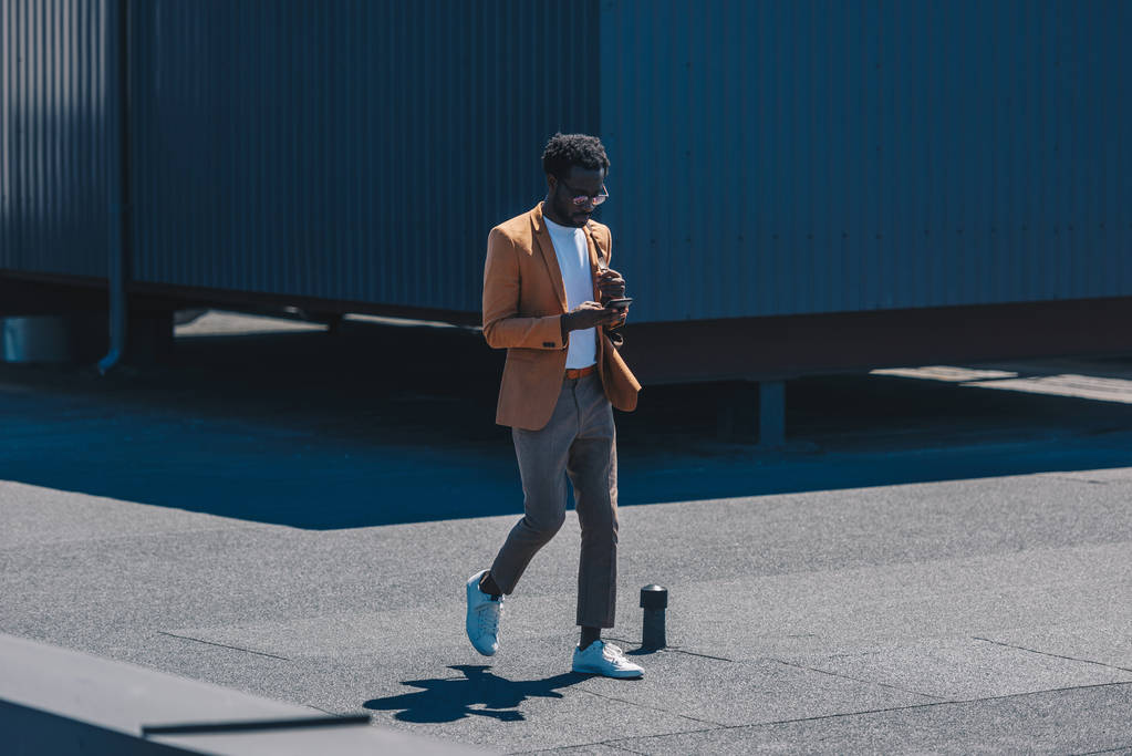 κομψό αφρικανικό Αμερικανό επιχειρηματία χρησιμοποιώντας φορητό υπολογιστή, ενώ περπατάει στην ταράτσα - Φωτογραφία, εικόνα