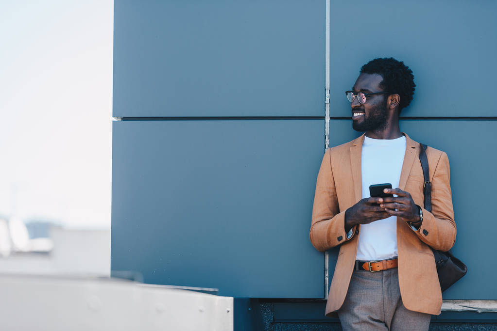 hymyilevä afrikkalainen amerikkalainen liikemies seisoo katolla ja katselee pois pitäen älypuhelin
 - Valokuva, kuva