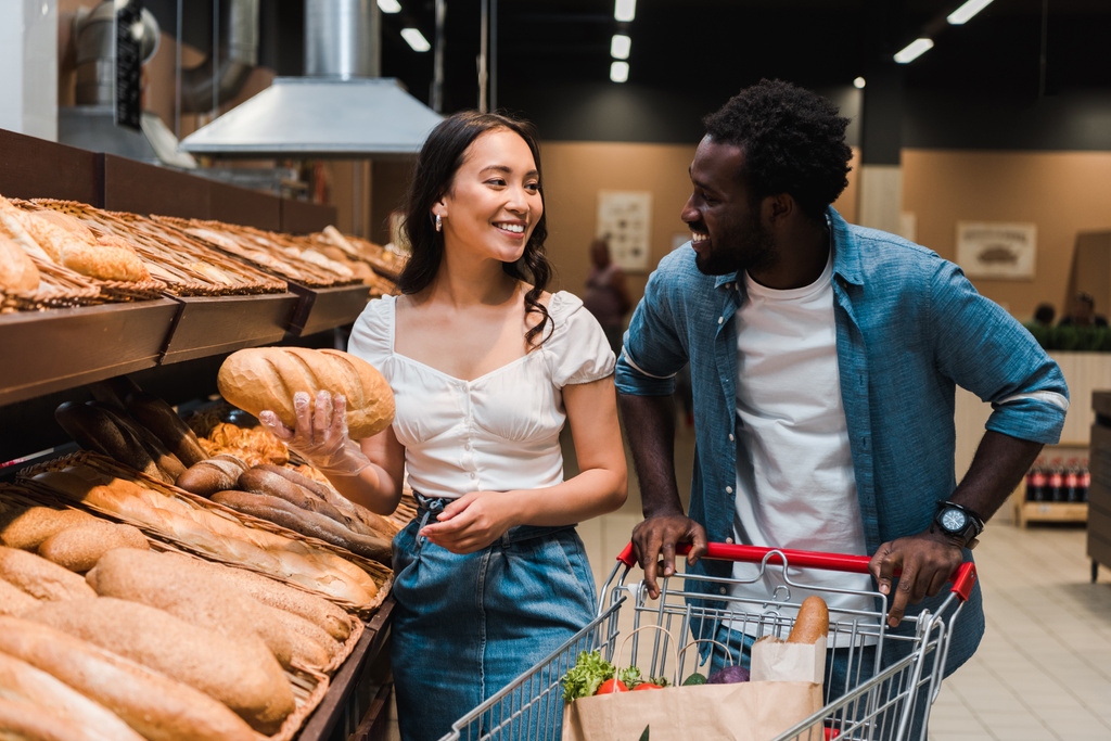 ショッピングカートで立っている陽気なアフリカ系アメリカ人男性の近くにパンを持つ幸せなアジアの女性  - 写真・画像