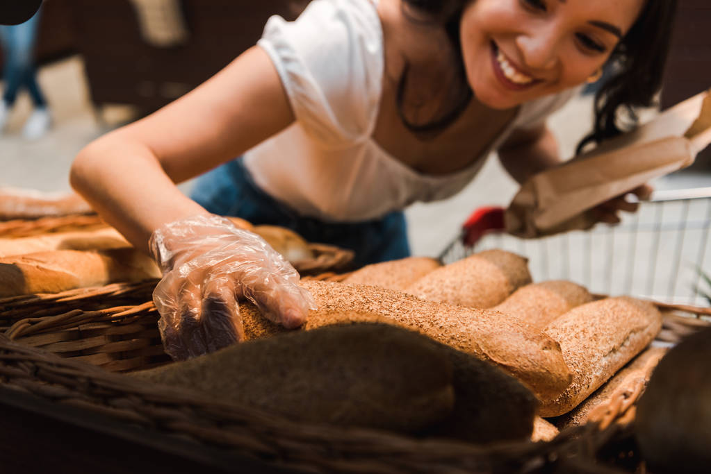 Ausgeschnittene Ansicht einer glücklichen Frau in Handschuhen in der Nähe von Brot im Supermarkt  - Foto, Bild