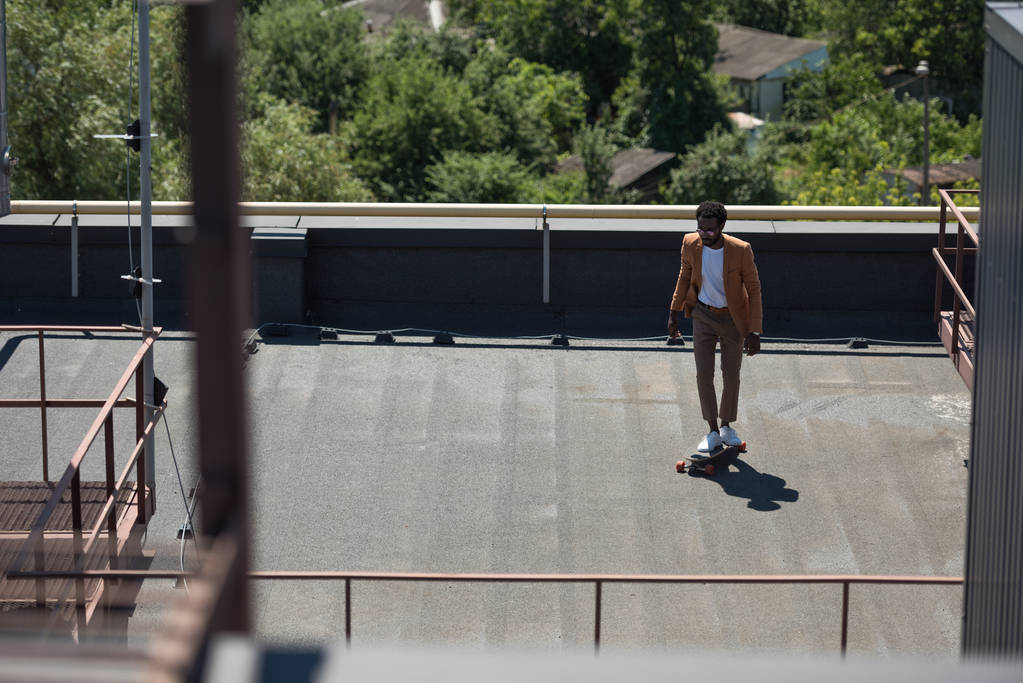 日当たりの良い屋上でロングボードに乗るスタイリッシュなアフリカ系アメリカ人ビジネスマンの選択的な焦点 - 写真・画像