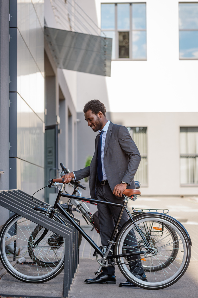 красивий афроамериканець бізнесмен посміхаючись, беручи велосипед з парковки  - Фото, зображення