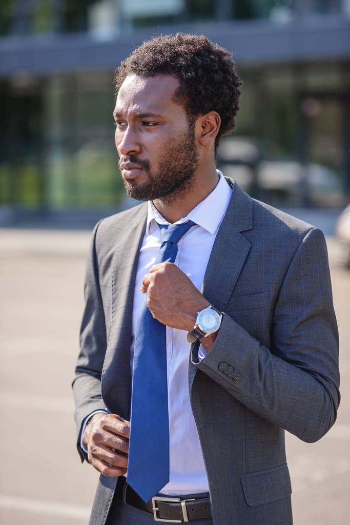 serious african american businessman touching tie and looking away on street - Photo, Image
