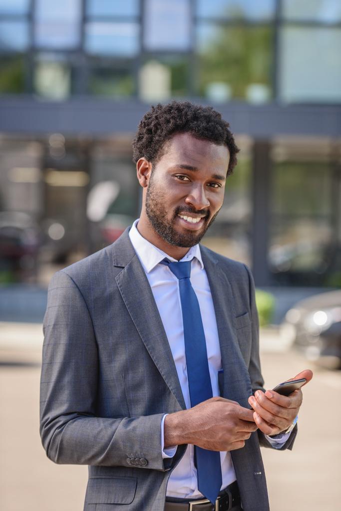 beau, souriant homme d'affaires afro-américain regardant la caméra tout en utilisant un smartphone
 - Photo, image