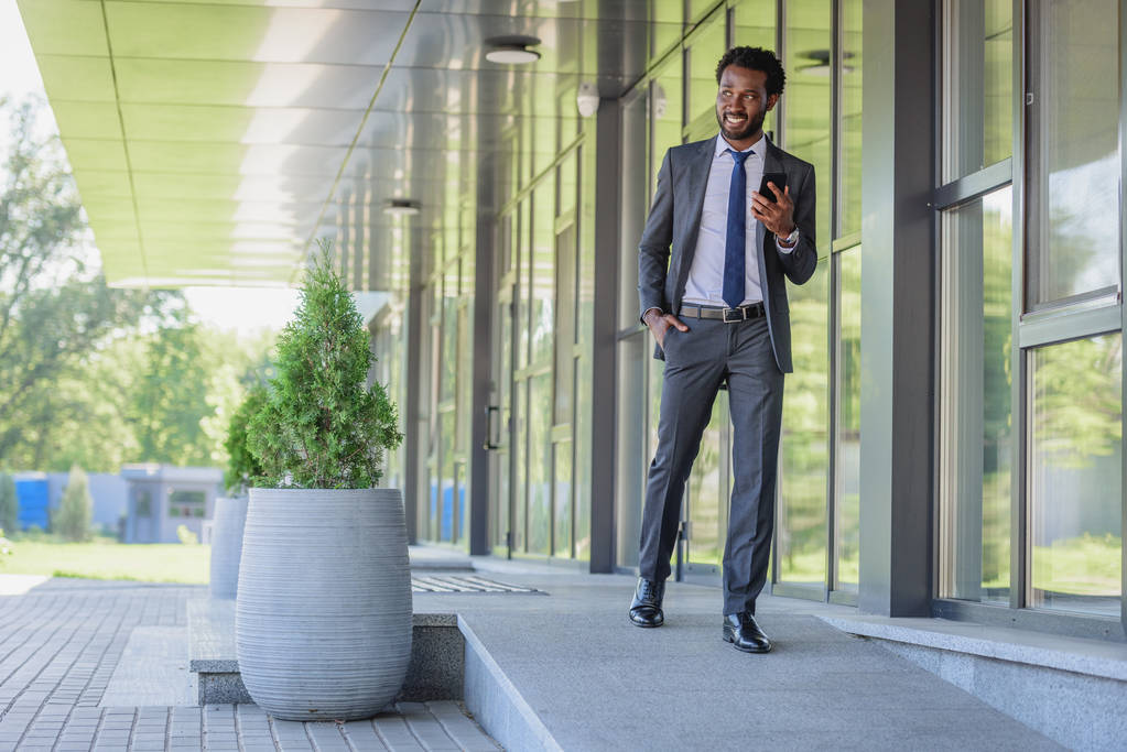 cheerful african american businessman holding smartphone while walking along office buiding - Photo, Image