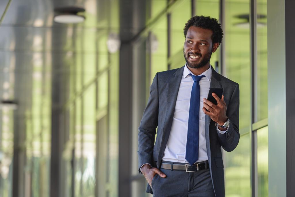 mosolygós afro-amerikai üzletember smartphone gyaloglás közelében irodaépület és kezében kéz a zsebben - Fotó, kép