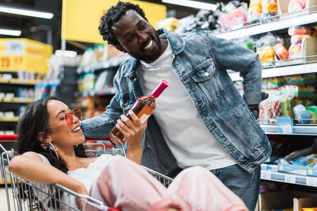 glückliche asiatische Mädchen mit Sonnenbrille hält Flasche mit Wein, während sie in einem Einkaufswagen in der Nähe afrikanisch-amerikanischer Mann sitzt  - Foto, Bild