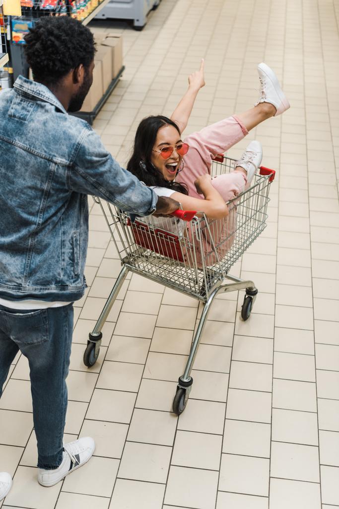 african american man walking with asian girl sitting in shopping cart and gesturing in supermarket  - Photo, Image
