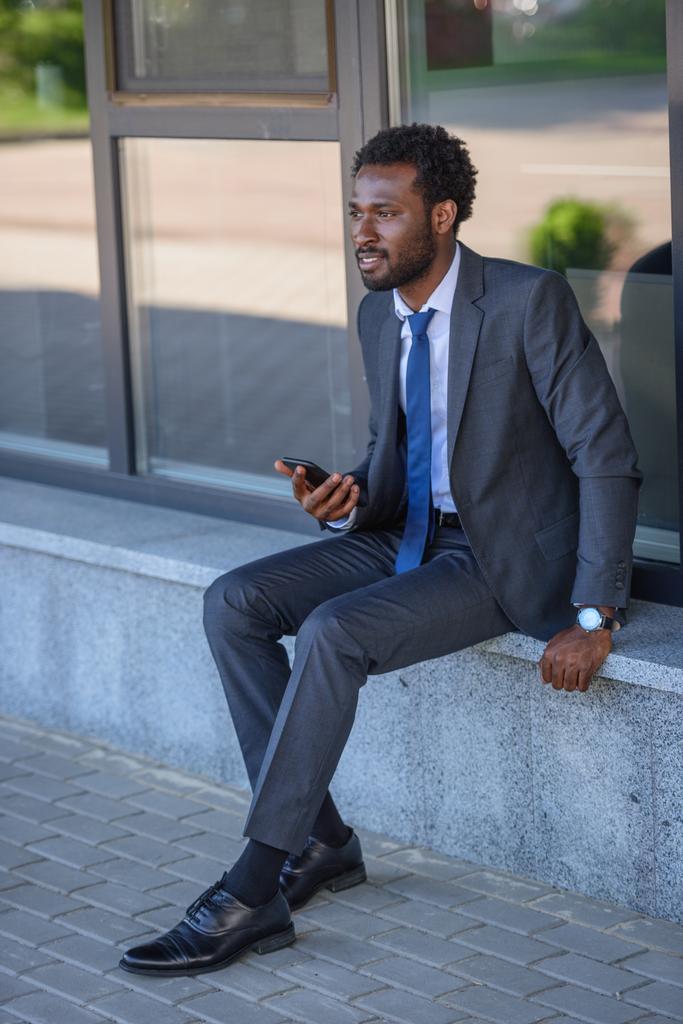 thoughtful african american businessman holding smartphone while sitting on parapet - Photo, Image