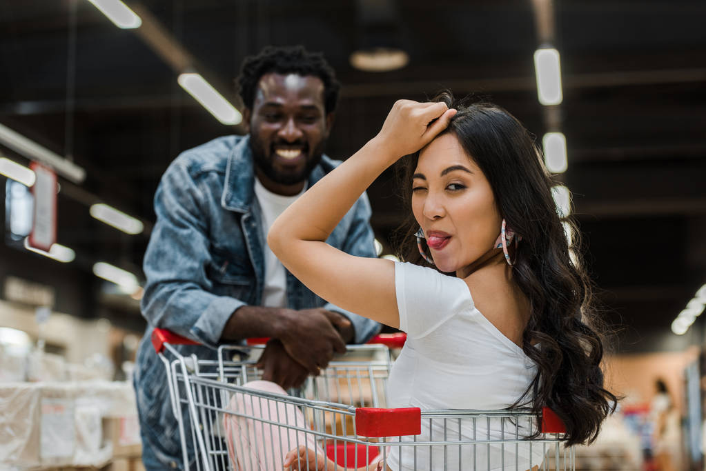 selective focus of cheerful asian girl sitting in shopping cart and showing tongue near african american man  - Photo, Image