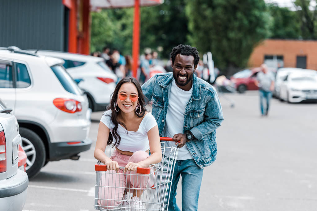 feliz homem americano africano andando enquanto alegre menina asiática em óculos de sol sentado no carrinho de compras
  - Foto, Imagem