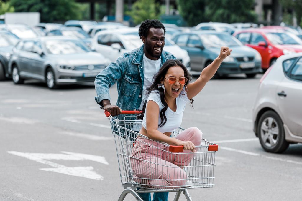Stylowy Asian Girl wskazując siedząc w wózek na zakupy w pobliżu African American Man i samochodów  - Zdjęcie, obraz