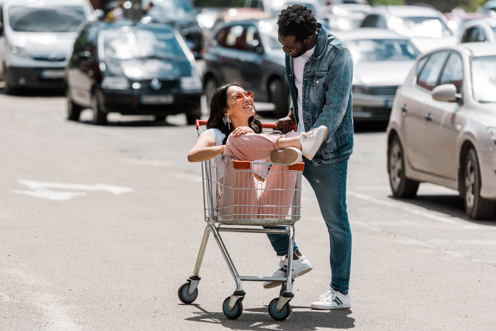 happy african american man standing near asian girl sitting in shopping cart in parking  - Photo, Image