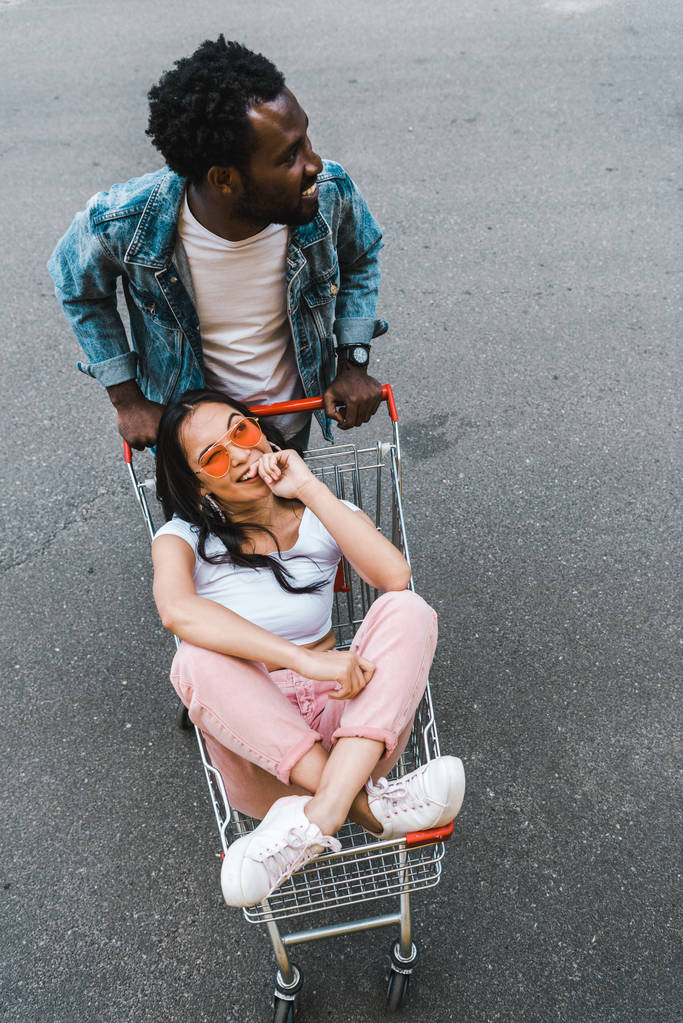 Widok z góry African American człowiek stoi w pobliżu szczęśliwy Asian dziewczyna siedzi w wózek na zakupy na zewnątrz  - Zdjęcie, obraz