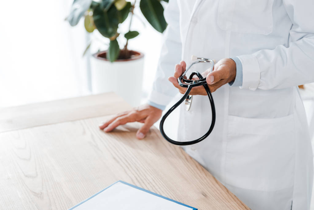 Ausgeschnittene Ansicht eines Arztes, der Stethoskop in der Nähe eines Holztisches hält  - Foto, Bild