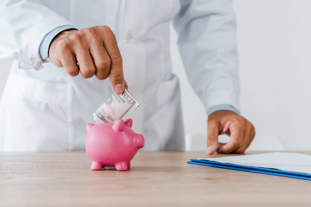 Ausgeschnittene Ansicht eines Arztes im weißen Kittel, der Dollarnoten in Sparschwein steckt  - Foto, Bild