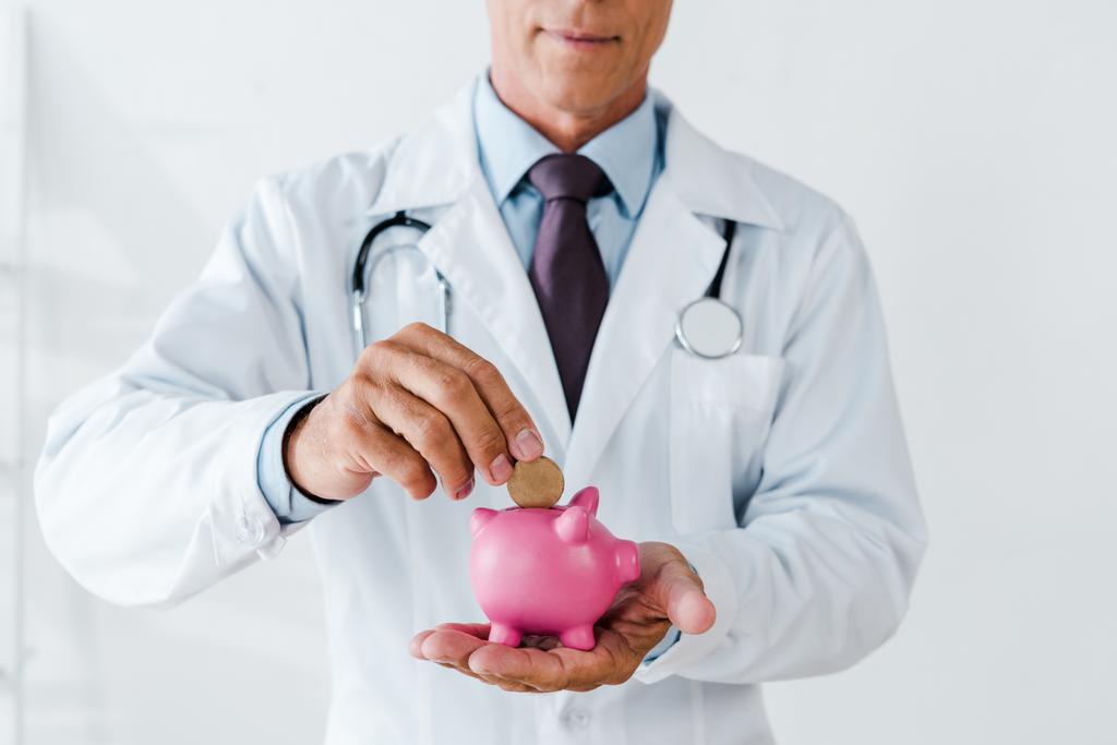 Ausgeschnittene Ansicht eines Arztes im weißen Kittel, der Münze in Sparschwein steckt  - Foto, Bild