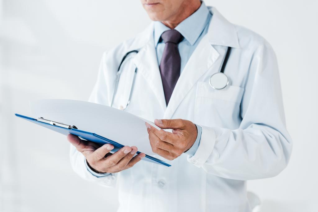 περικομμένη προβολή του γιατρού σε λευκό παλτό κρατώντας σελίδα και πρόχειρο στην κλινική  - Φωτογραφία, εικόνα