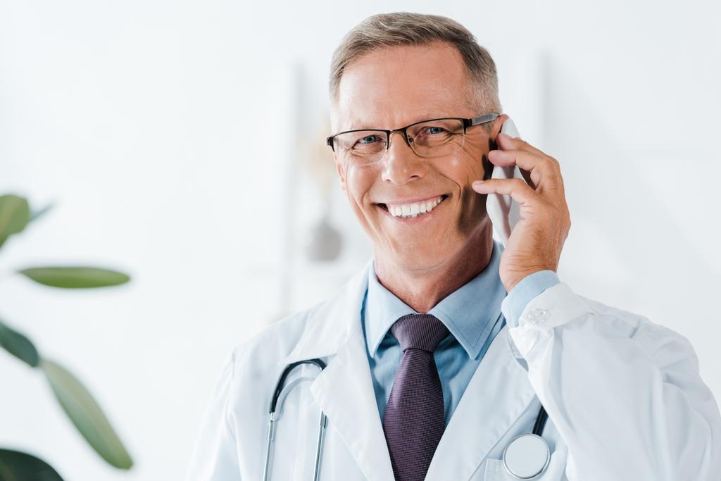 ευτυχισμένος γιατρός κοιτάζοντας την κάμερα και μιλάει στο smartphone στην κλινική  - Φωτογραφία, εικόνα