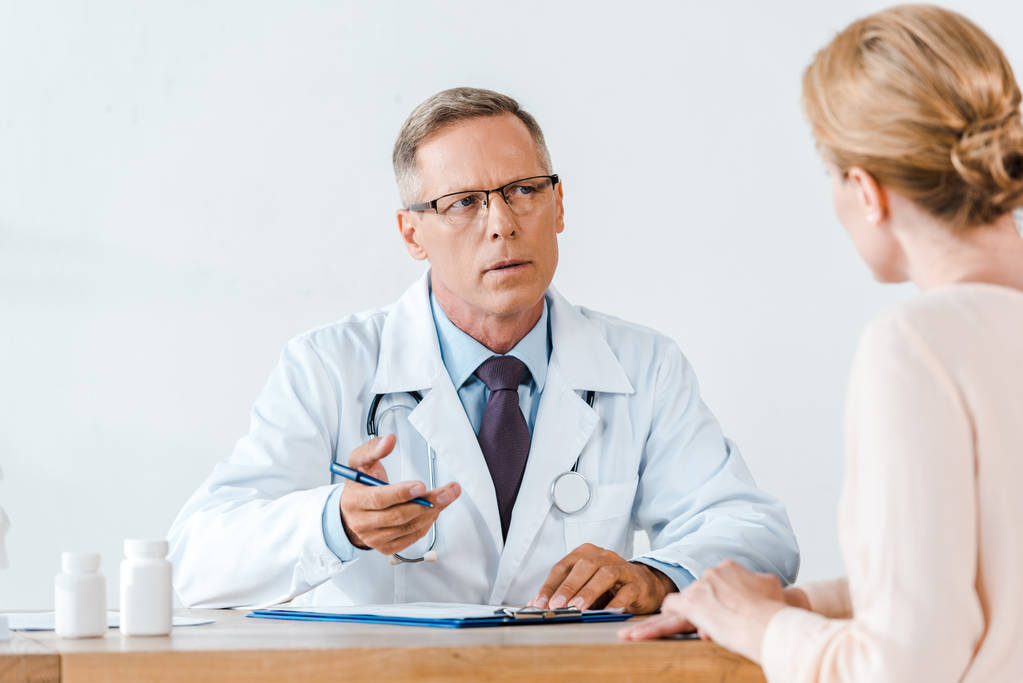 selective focus of doctor in white coat looking at woman and gesturing while sitting near table  - Photo, Image