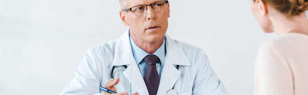 panoramatický záběr doktora v bílém plášti, který se dívá na ženu a gestikulovat na klinice  - Fotografie, Obrázek