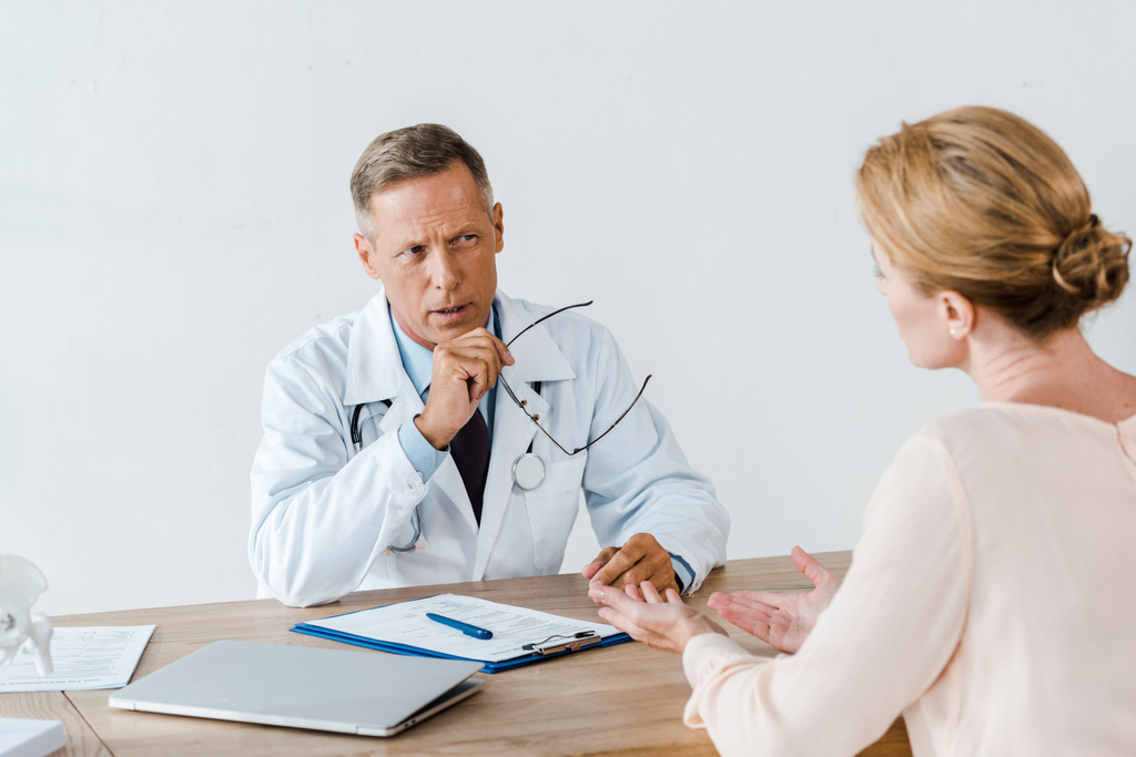 селективный фокус врача в белом халате, смотрящего на женщину и держащего очки в больнице
 - Фото, изображение