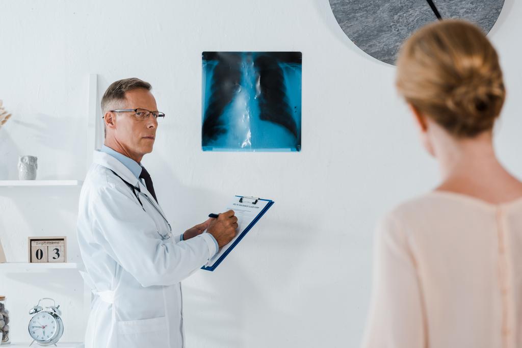 selective focus of doctor in glasses writing diagnoses on clipboard near x-ray and looking at woman in clinic  - Photo, Image