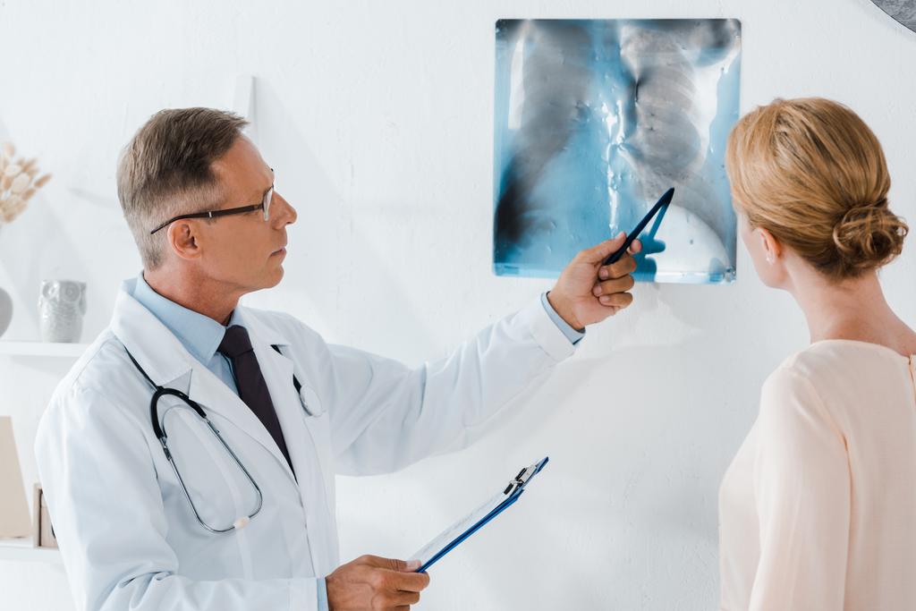 лікар в окулярах тримає ручку біля рентгенівського випромінювання і жінку в клініці
  - Фото, зображення