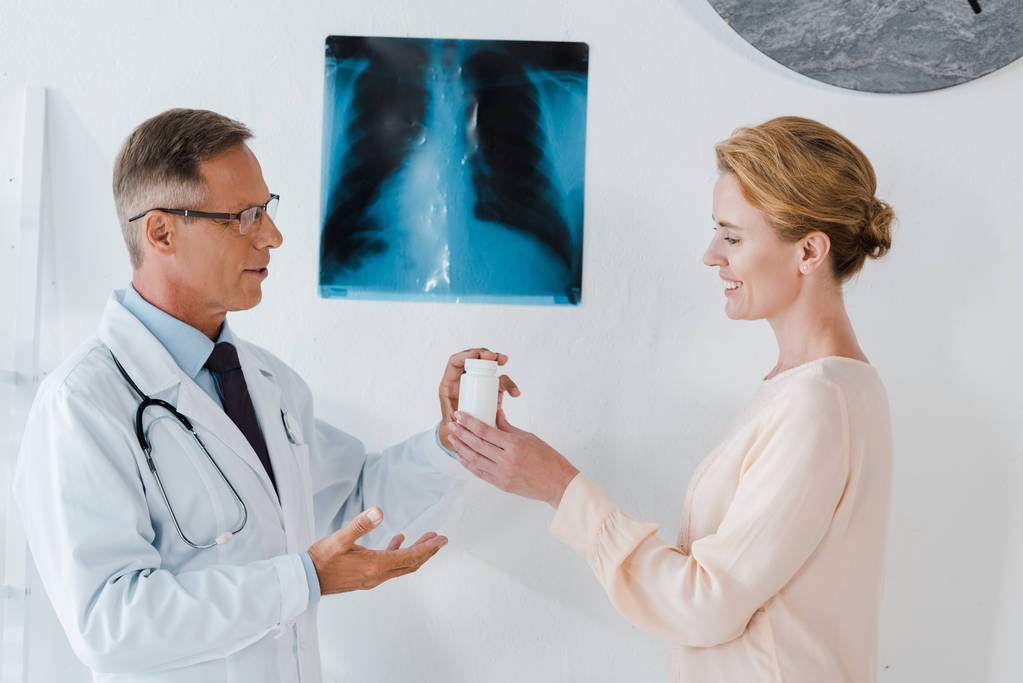 schöner Arzt mit Brille, der neben dem Röntgenbild steht und der fröhlichen Frau eine Flasche gibt  - Foto, Bild