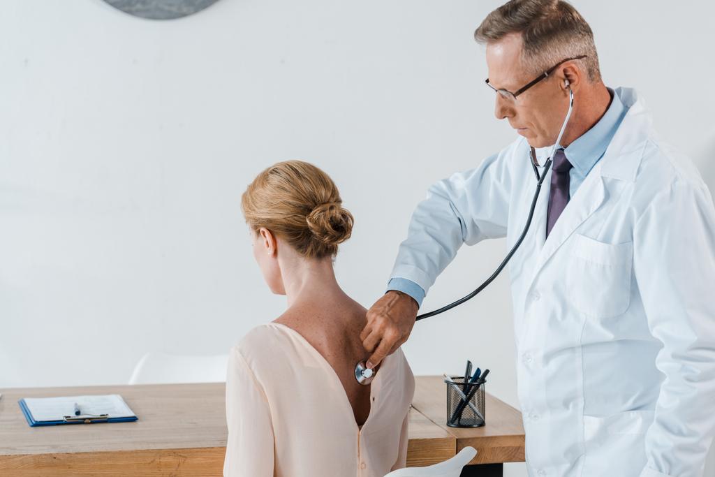 arts in glazen met behulp van stethoscoop tijdens het onderzoeken van de vrouw in de kliniek  - Foto, afbeelding