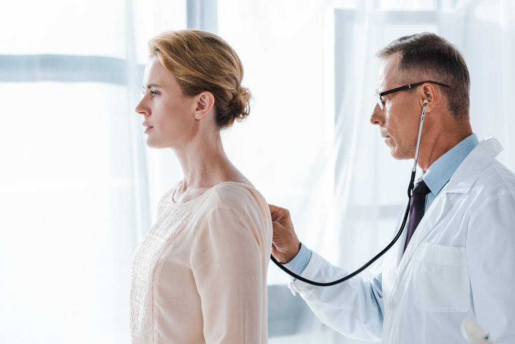 Widok z boku lekarza w okularach za pomocą stetoskop podczas badania atrakcyjny pacjenta w klinice  - Zdjęcie, obraz