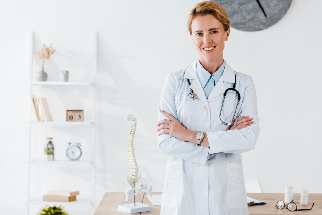 aantrekkelijke en positieve arts staande met gekruiste armen in de kliniek  - Foto, afbeelding