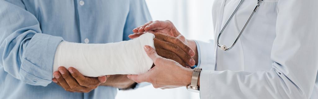 панорамный снимок врача, трогающего сломанную руку человека в клинике
  - Фото, изображение