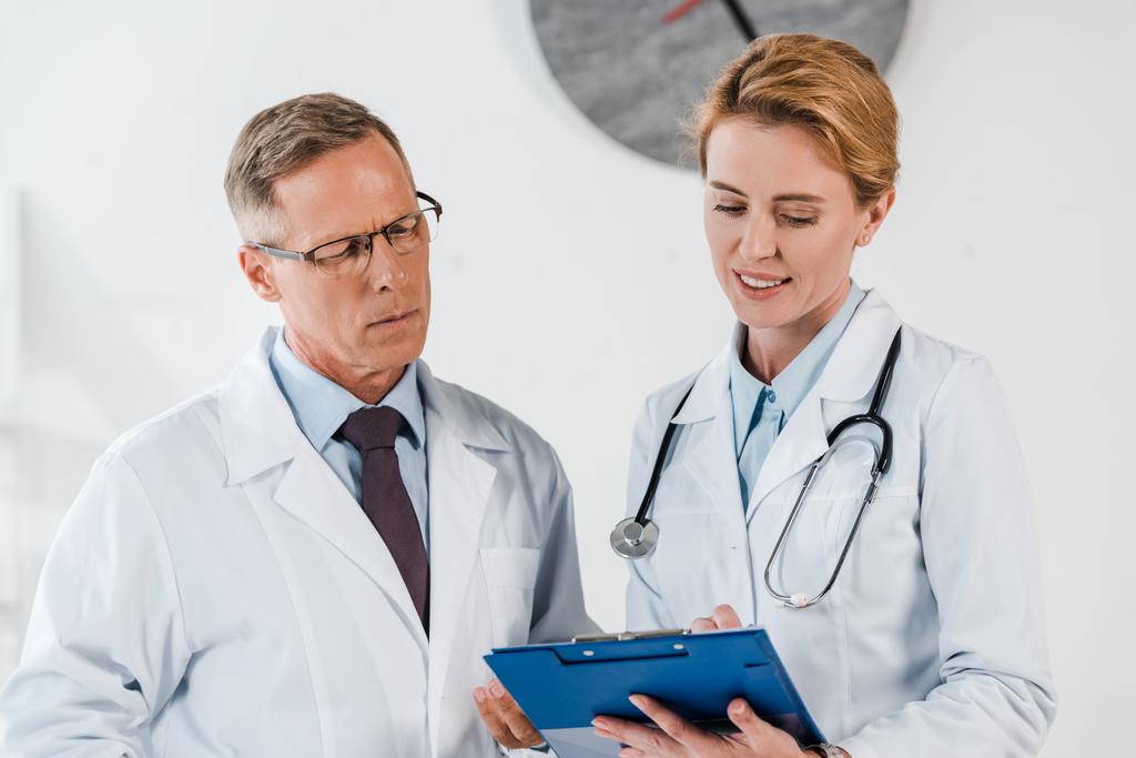 handsome doctor standing near happy coworker and looking at clipboard  - Photo, Image