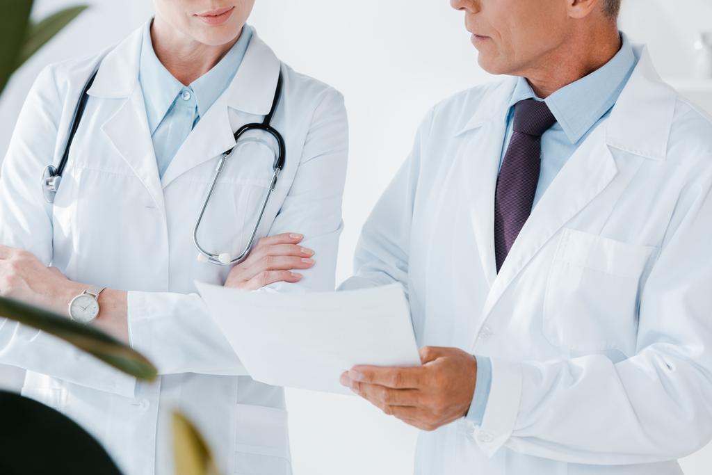 vista cortada de médico segurando papel em branco perto colega com braços cruzados
  - Foto, Imagem