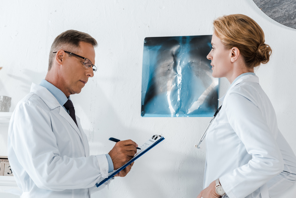 handsome man in glasses writing while holding clipboard near x-ray and attractive coworker  - Photo, Image