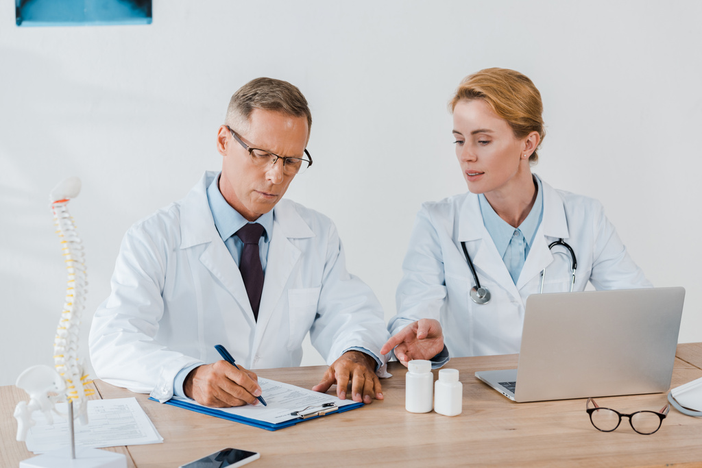 привлекательный доктор, указывающий пальцем на ноутбук и коллега в очках, пишущий диагноз
  - Фото, изображение