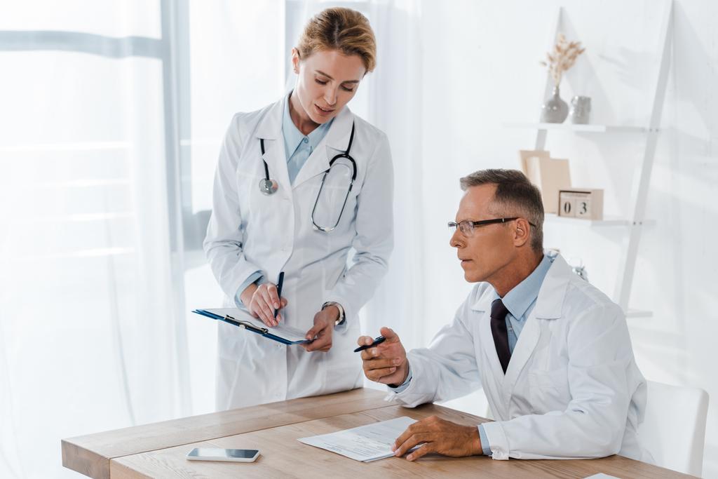 привлекательный врач держит планшет и ручку, стоя рядом с коллегой и столом
  - Фото, изображение