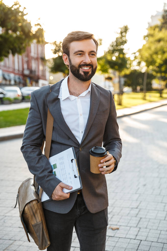 Muotokuva onnellinen aikuinen liikemies virallisessa puku tilalla takeaway kahvia kävellessään läpi kaupungin katu
 - Valokuva, kuva