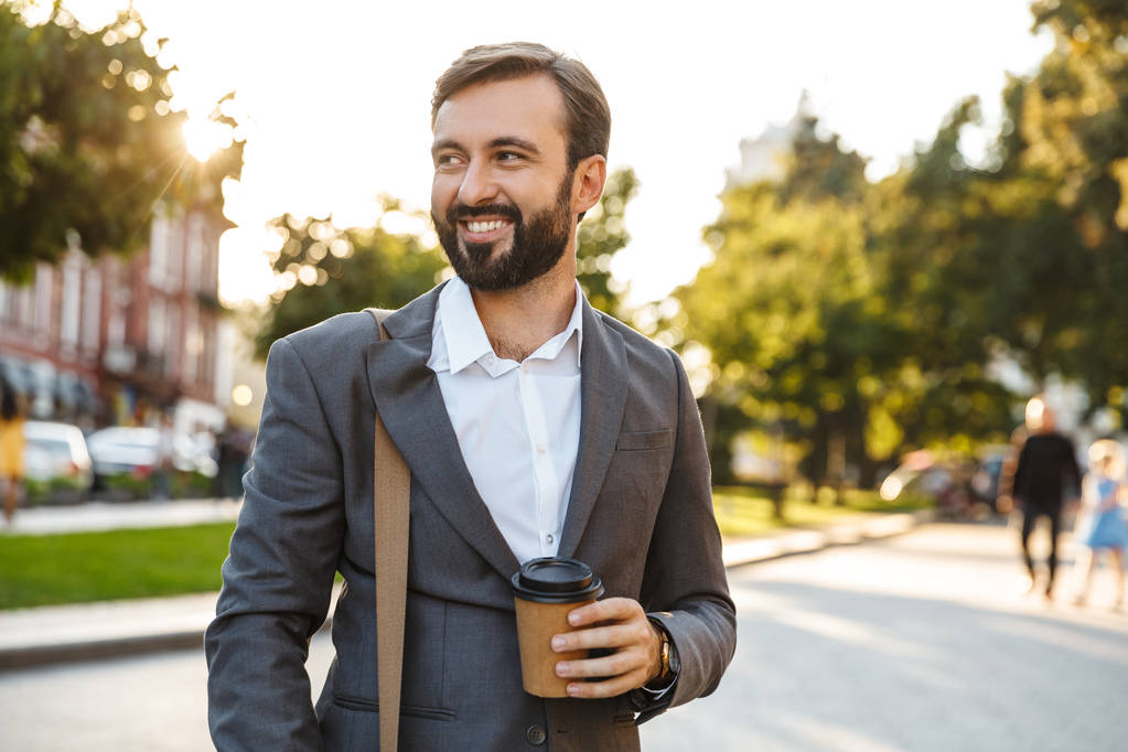 Πορτραίτο του χαμογελαστή ενήλικα επιχειρηματία σε επίσημο κοστούμι κρατώντας καφέ σε πακέτο ενώ περπατάει μέσα από την οδό της πόλης - Φωτογραφία, εικόνα