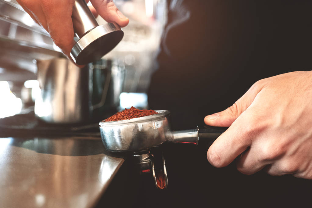 バリスタは、片手に挽いたコーヒーを持つコーヒーホルダーを保持し、カフェでアメリカーノを準備する別の近くのプロのコーヒーマシンで改ざんクローズアップ - 写真・画像