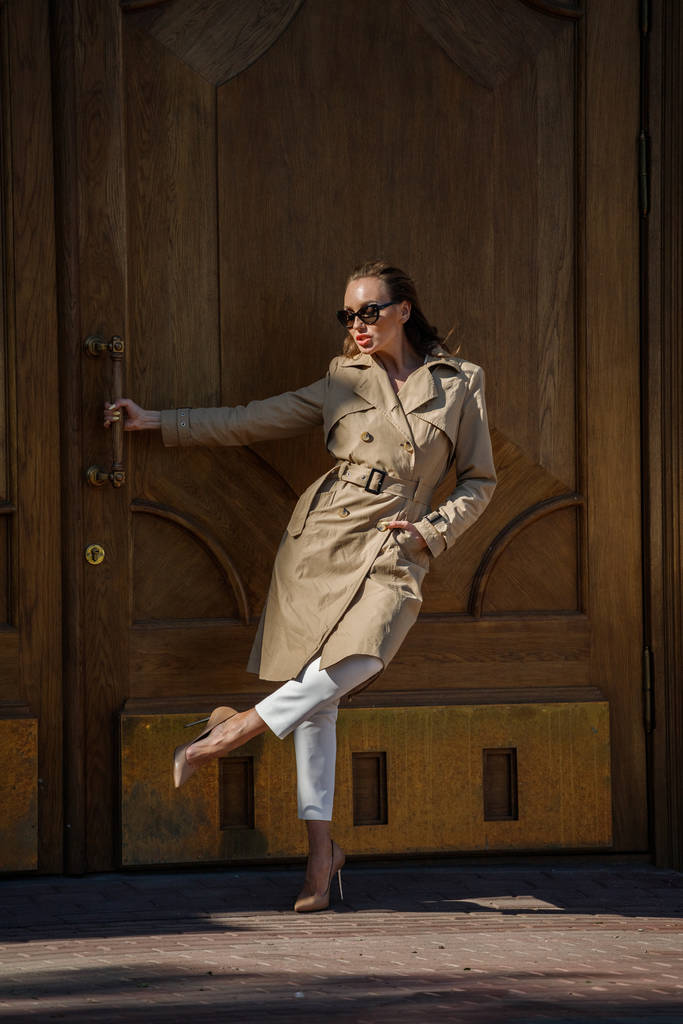 Blonďatá žena stojící venku ve dne. Portrét stylového modelu s béžovým croakem, bílými kalhotami a slunečními brýlemi  - Fotografie, Obrázek