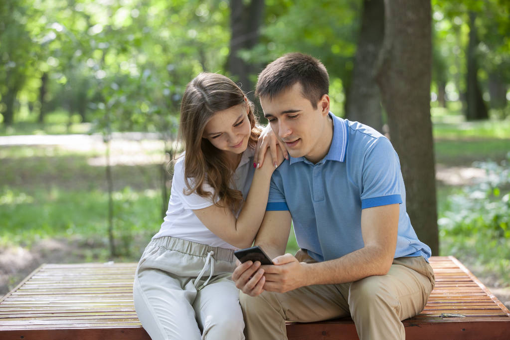 Pareja compartiendo medios en un teléfono inteligente sentado en un banco en un parque
. - Foto, imagen