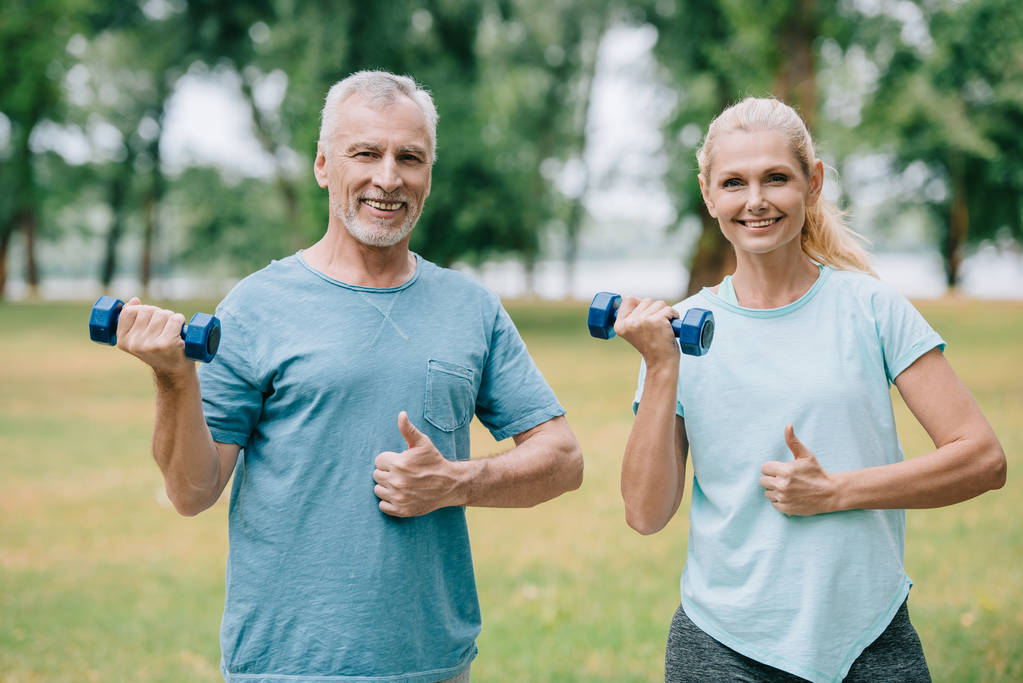 smiling mature sportsman and sportswoman holding barbells and showing thumbs up while smiling at camera - Photo, Image
