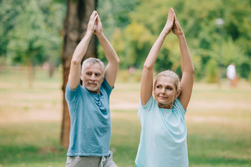mature sportsman and sportswoman stretching with raised hands while training in park - Photo, Image