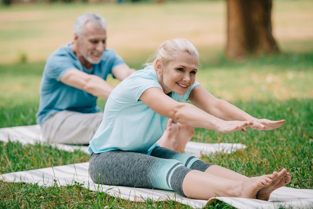 selective focus of smiling, mature man and woman sitting in stretching poses while practicing yoga in park - Photo, Image
