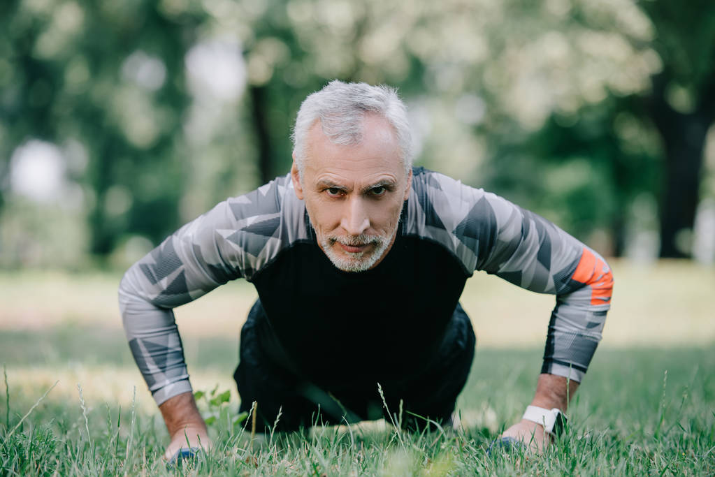 confident mature sportsman doing push ups with barbells on lawn in park - Photo, Image