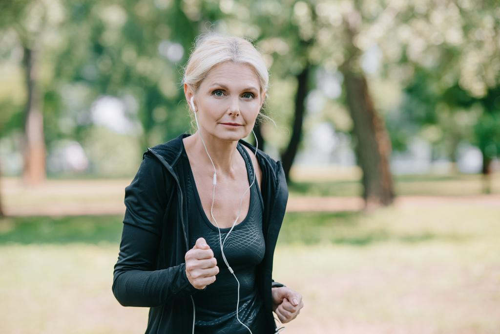 beautiful mature sportswoman listening music in earphones while jogging in park - Photo, Image